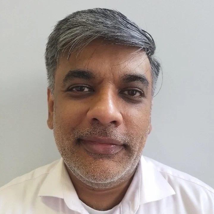 A headshot of a middle-aged man with graying hair and a beard, wearing a white collared shirt.