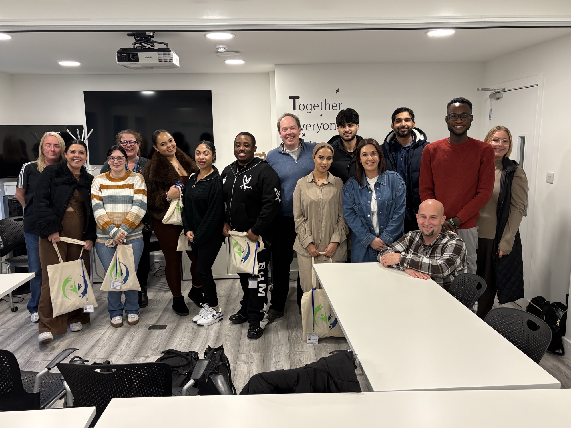 A diverse group of 14 people are posing for a group photo in a bright room with a projector and large screens. They are holding tote bags.